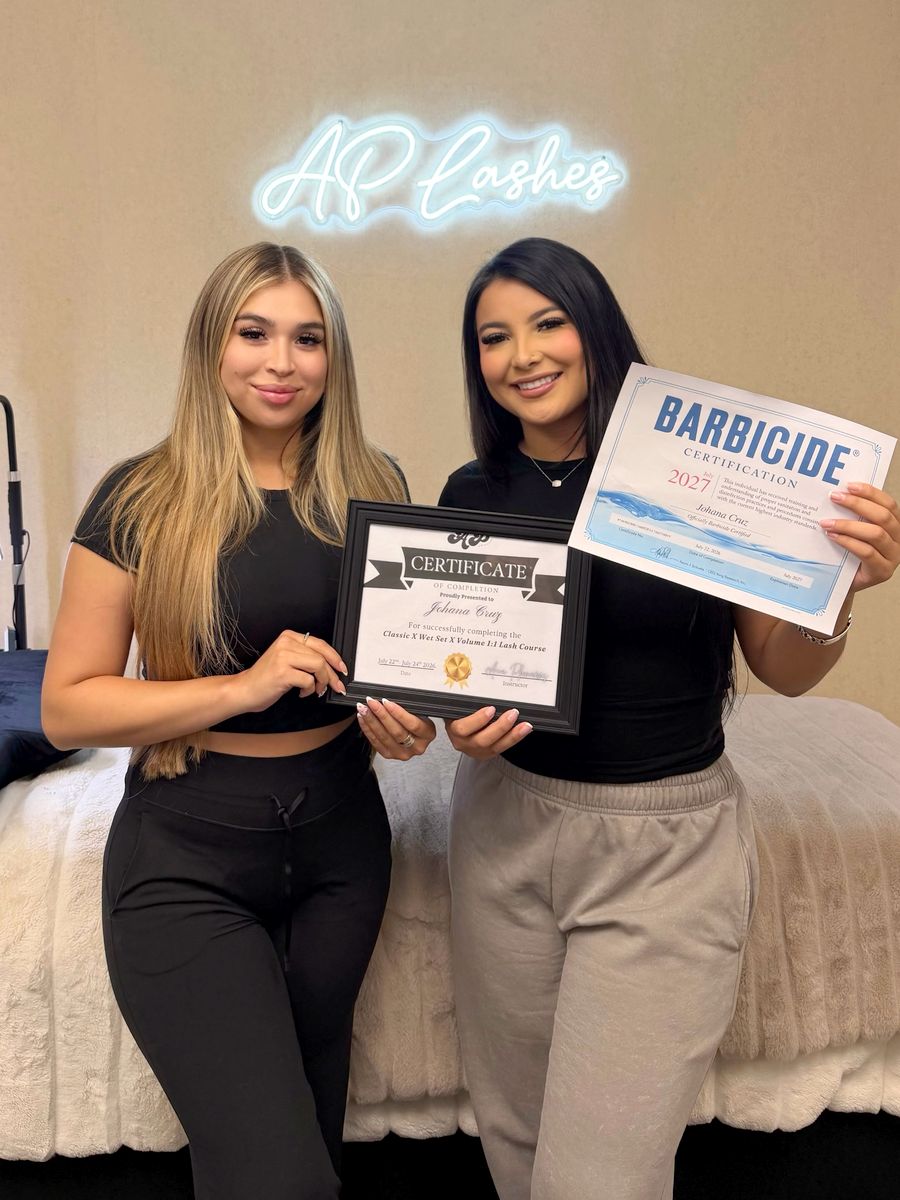 Two graduates with certificates under the AP Lashes neon sign